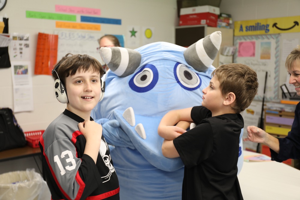 Two young male students hug a blue monster mascot from learning software they use in class