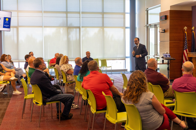 A man in a suit speaks to a group of 20 adults sitting in green chairs.