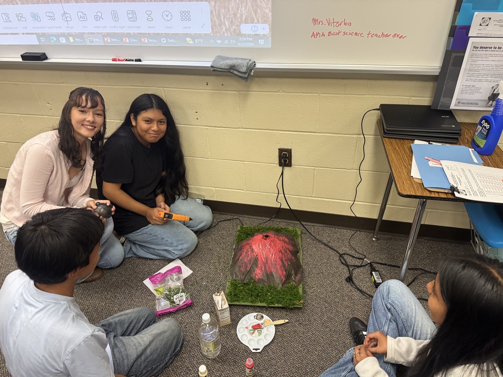 Students are sitting on the floor in a classroom testing their volcano models.