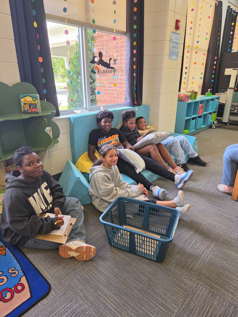 Several students sit in a classroom, with one girl seated on a blue storage basket. Two boys sit on a bench behind them.