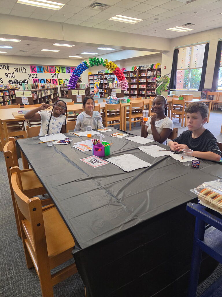 Four children at a desk with a black table cover, surrounded by chairs and bookshelves.