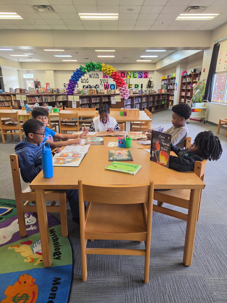 Group of children seated around a table in a library. Books, a backpack, and a water bottle on table.