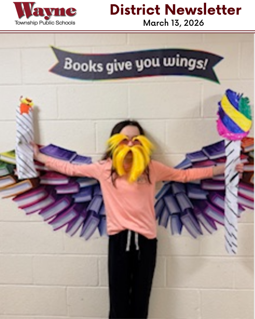 Child in pink shirt, yellow wig, and beard stands in front of wall with banner and wings.