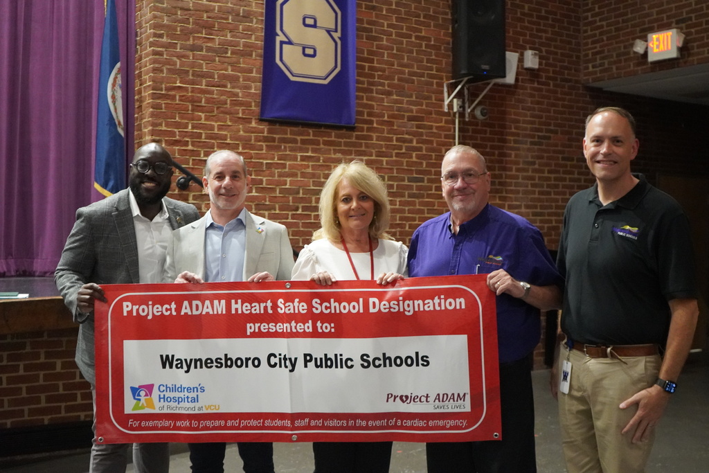 A group of school health professionals and community partners stands together at a Waynesboro School Board meeting, holding a banner that reads, “Project ADAM Heart Safe School Designation — Waynesboro Public Schools.