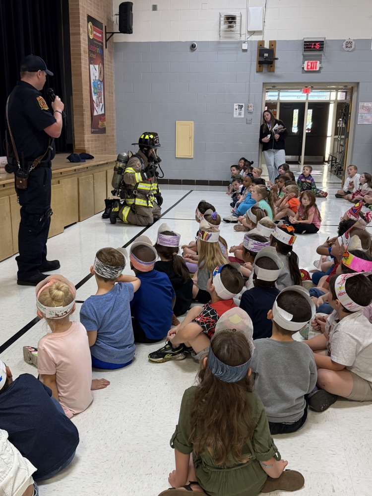 a group of students sitting and listening to a fireman talk. There is also a firefighter dressed in their gear kneeling on the ground