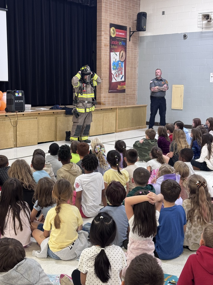 A group of students watching and listening to the firefighter speak. There is also a firefighter with their gear on speaking to the students
