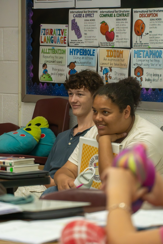 Two high school student, one male and one female smiling while watching the teacher teach her students.