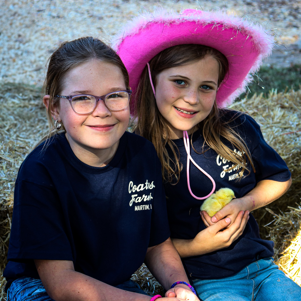 2 girls at farm day - one wearing a pink furry cowgirl hat and holding a baby chicken, the other smiling