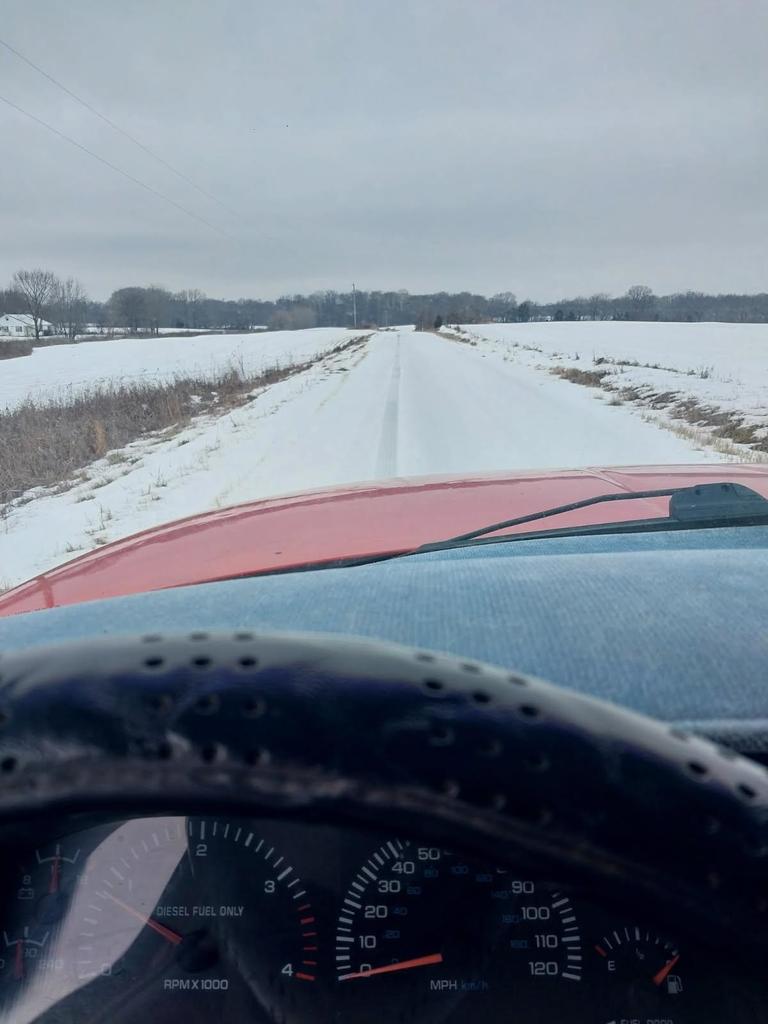 View from inside a vehicle looking down a rural road covered in snow, with tire tracks visible but icy conditions remaining on the roadway. Snow-covered fields line both sides of the road under an overcast sky