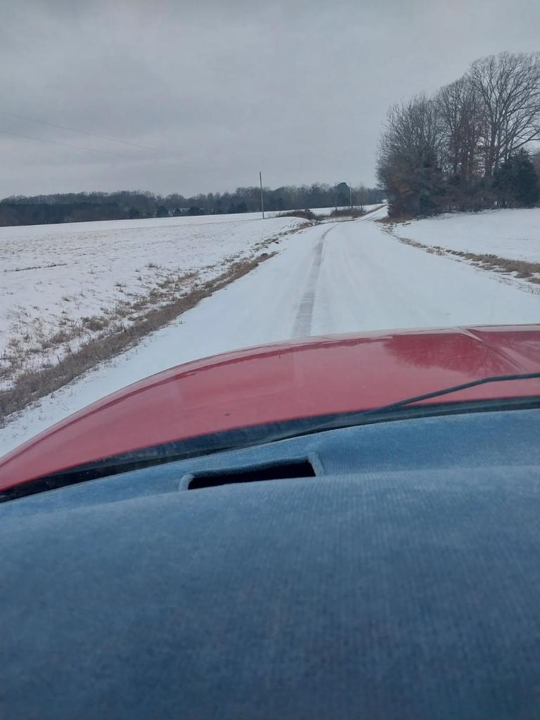 View from inside a vehicle looking down a rural road covered in snow, with tire tracks visible but icy conditions remaining on the roadway. Snow-covered fields line both sides of the road under an overcast sky