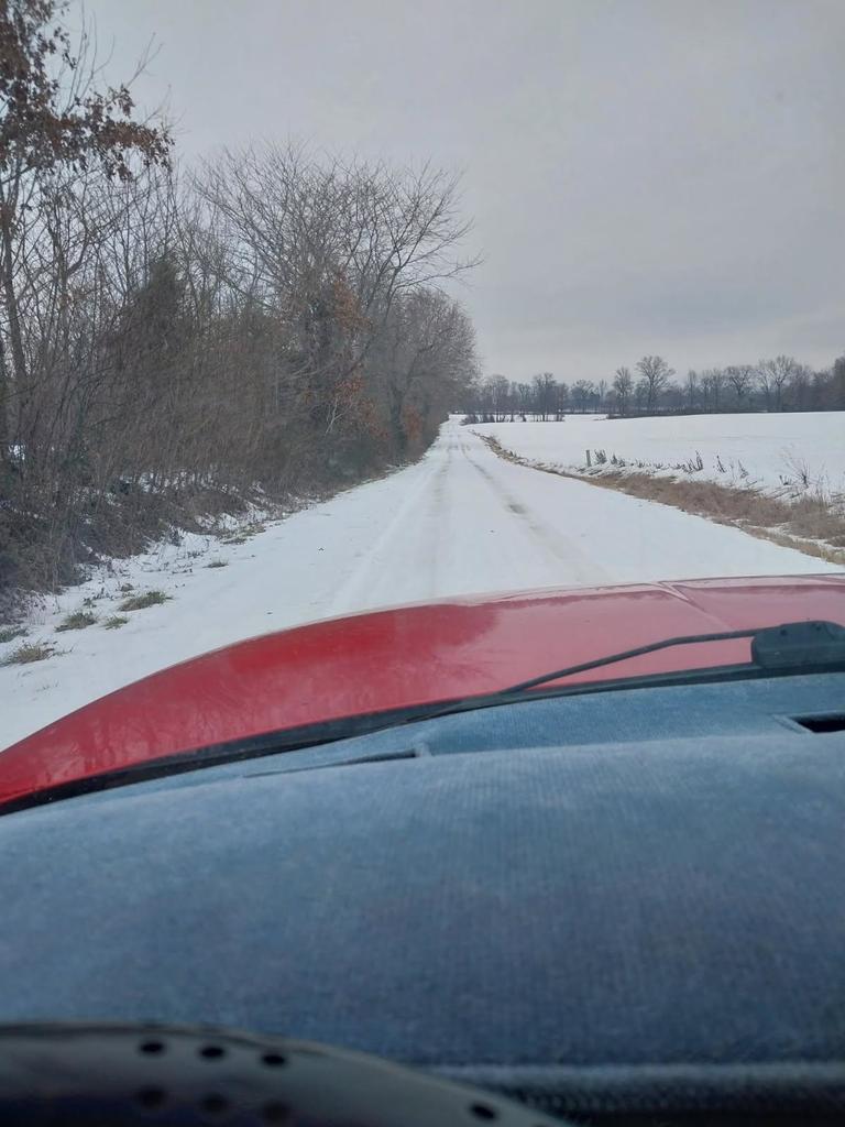 View from inside a vehicle looking down a rural road covered in snow, with tire tracks visible but icy conditions remaining on the roadway. Snow-covered fields line both sides of the road under an overcast sky
