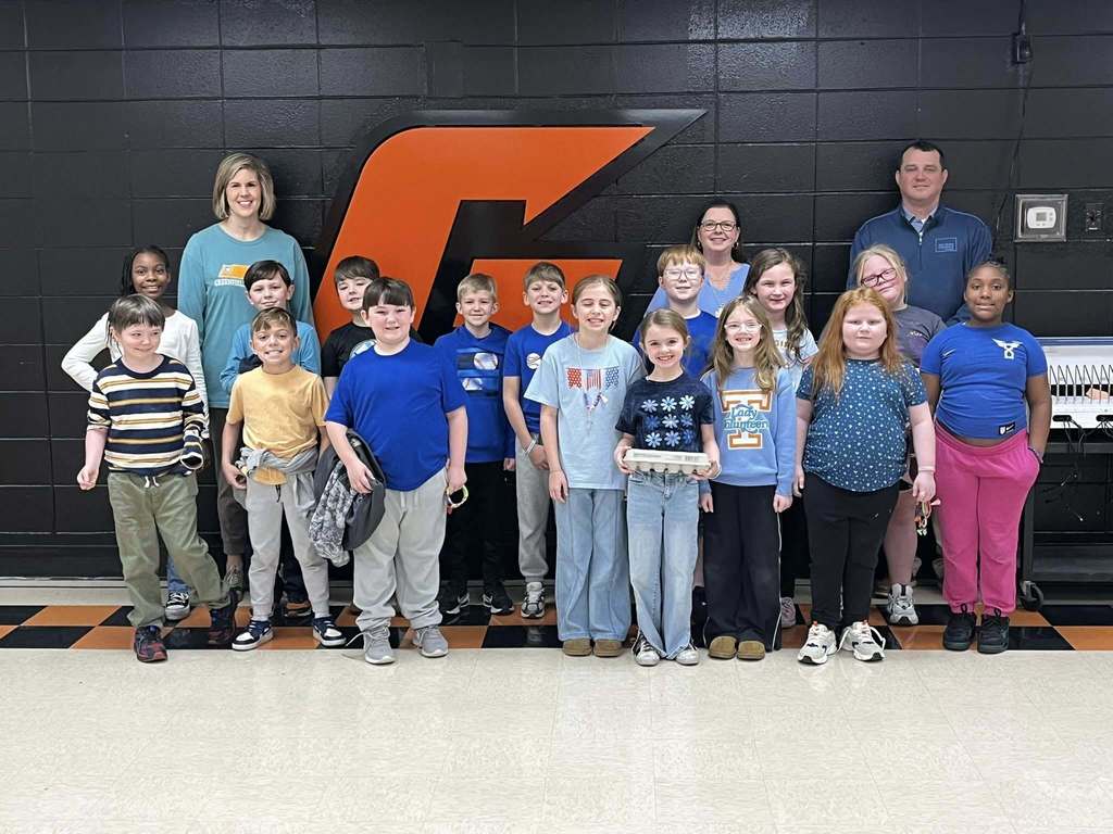 Elementary students and teachers pose in a school hallway in front of a large orange letter on a black wall. One student holds an egg carton while others stand smiling in rows.