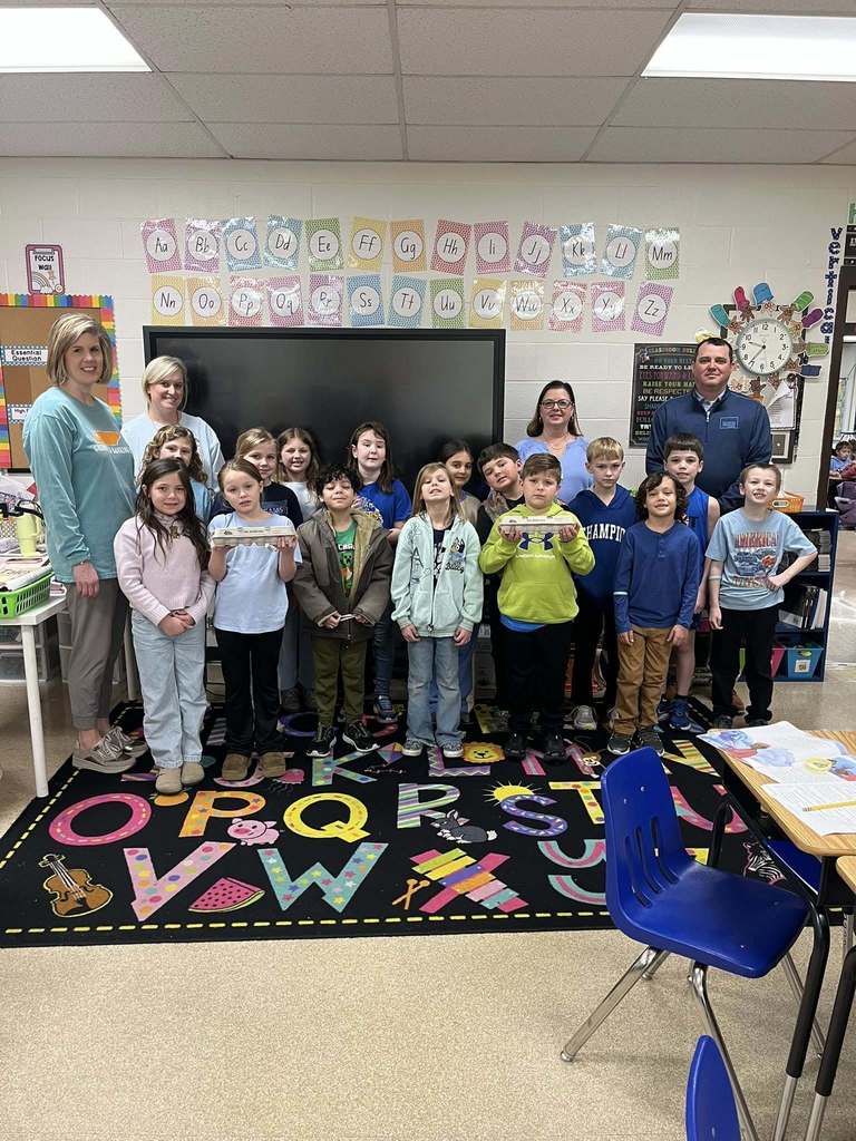 Students and teachers stand together in a bright classroom decorated with alphabet letters and colorful rugs. Several students hold egg cartons as part of a hands-on agriculture lesson.