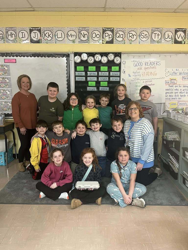 A class of elementary students and two teachers pose in a classroom with reading and phonics posters on the wall. One student sits in front holding an egg carton while classmates stand closely together.