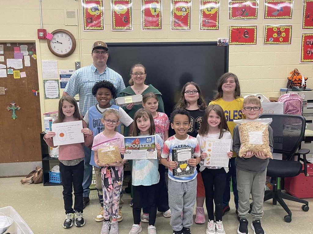 Students and teachers stand together in a bright classroom decorated with alphabet letters and colorful rugs. Several students hold egg cartons as part of a hands-on agriculture lesson.