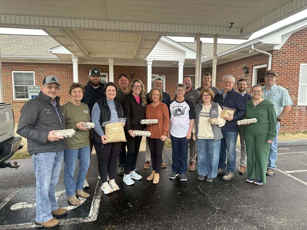 A group of adults stand outside a school building holding cartons of eggs and bags of chick feed. The group smiles for a photo under a covered entrance on a rainy day.