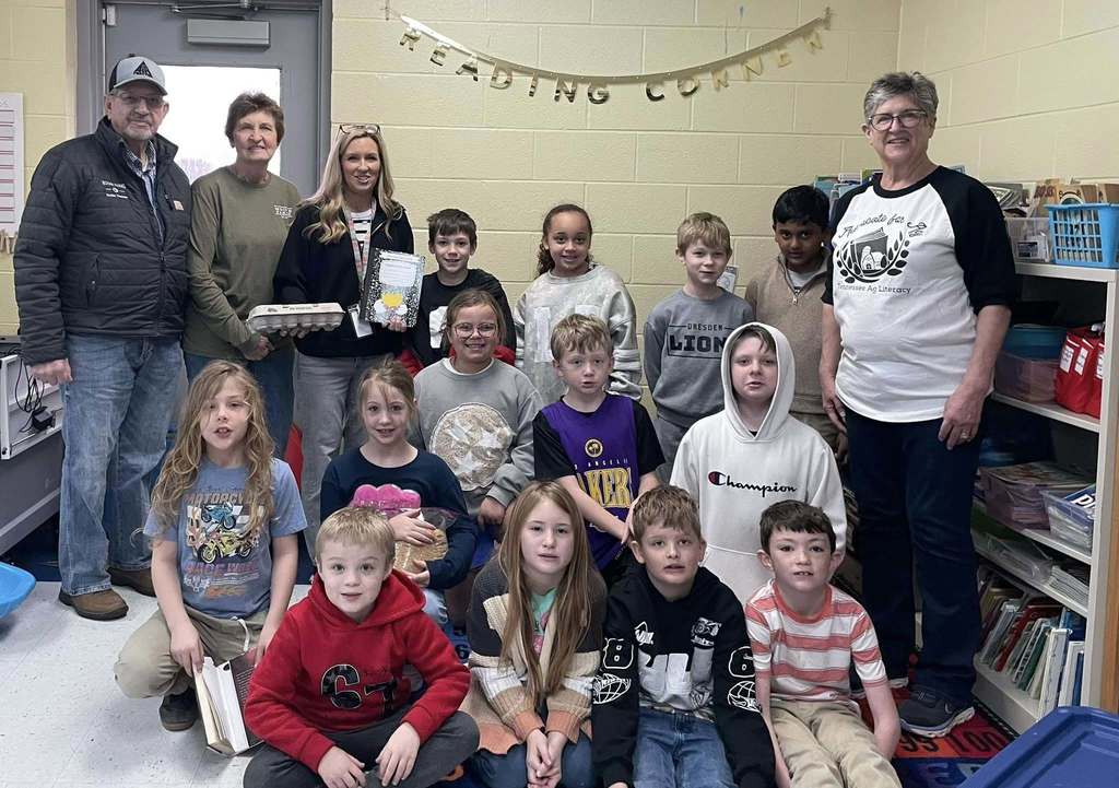 Elementary students pose in a classroom with several adult volunteers, holding eggs, notebooks, and poultry-related materials. A banner reading “Reading Corner” hangs on the wall behind them.