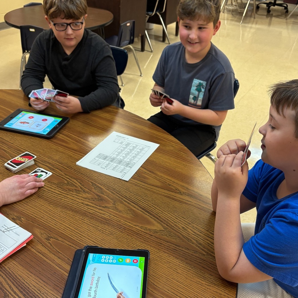 Three elementary students sit at a table playing UNO cards while following along with learning activities on tablets during a small group instructional session.