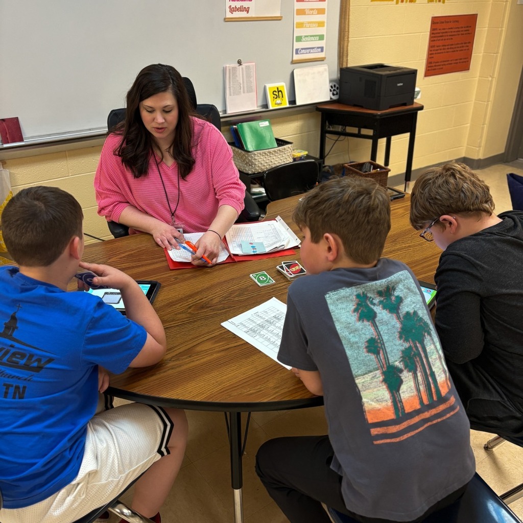 A teacher leads a small group of students at a round table, reviewing materials and guiding them through an interactive lesson using cards and tablets.