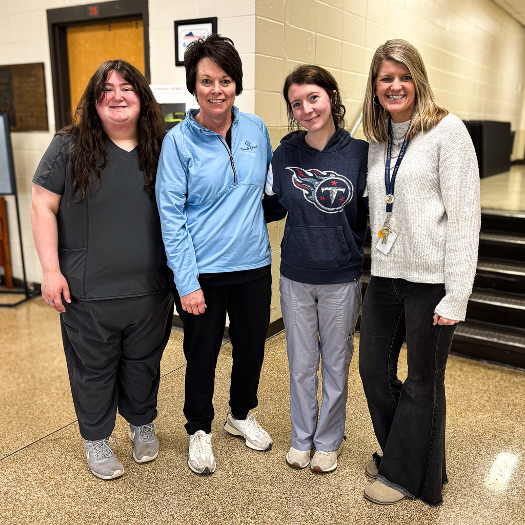 Four educators stand together in a school hallway, smiling for a photo. They are dressed casually in comfortable, school-appropriate clothing, including scrubs, athletic wear, and a sweater with a lanyard. The group poses shoulder-to-shoulder near a classroom door and stairwell, creating a warm, friendly team moment.