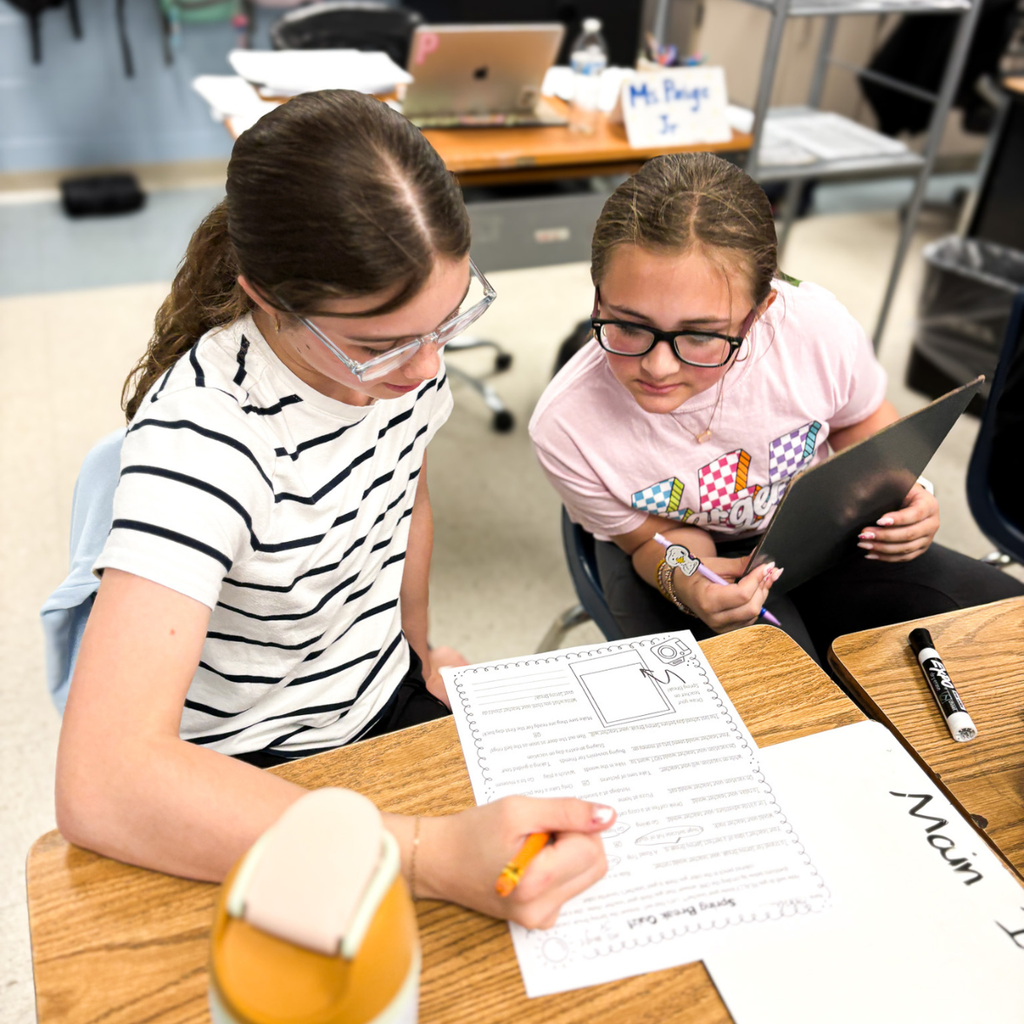 Two students collaborate at a desk, reading a worksheet and discussing answers about the main idea.