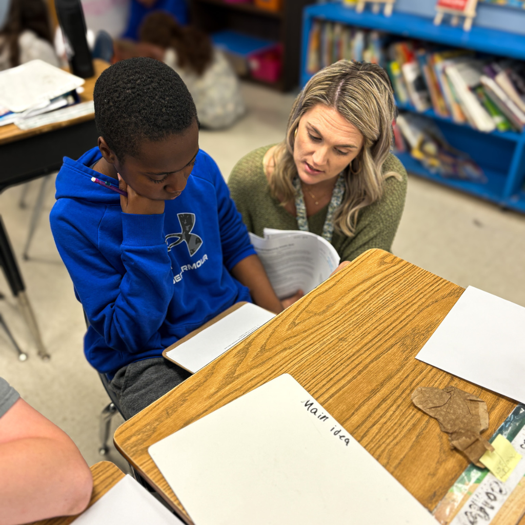 Paige Hatchel kneels beside a student, offering one-on-one support while reviewing a worksheet about main ideas.