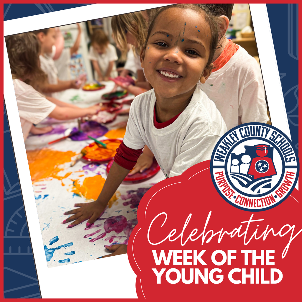 Children in a classroom paint on a large table. One child smiles with paint on their face.