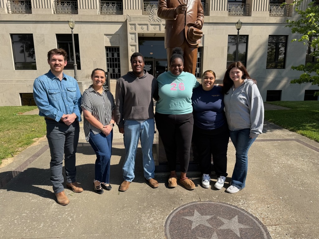 Six individuals stand together in front of a large statue, on a concrete area with a circular emblem.