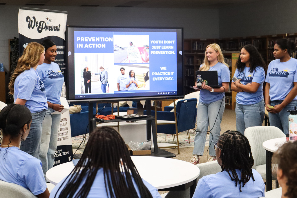 A group of women in blue shirts stands in front of a screen in a room with tables and chairs.