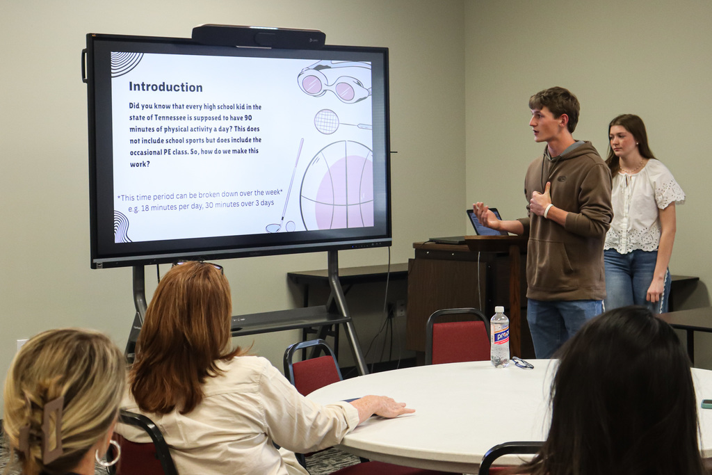 A man stands at a podium in a classroom, addressing a group of people seated around a table.