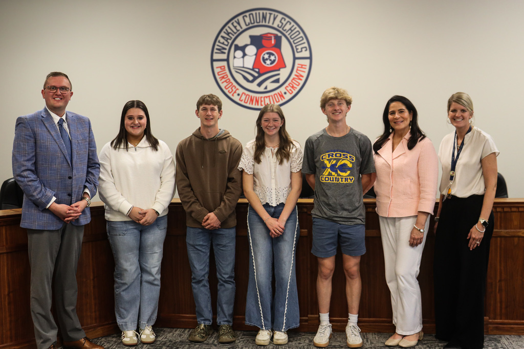 A group of people stands behind a desk, smiling, with a school logo on the wall.