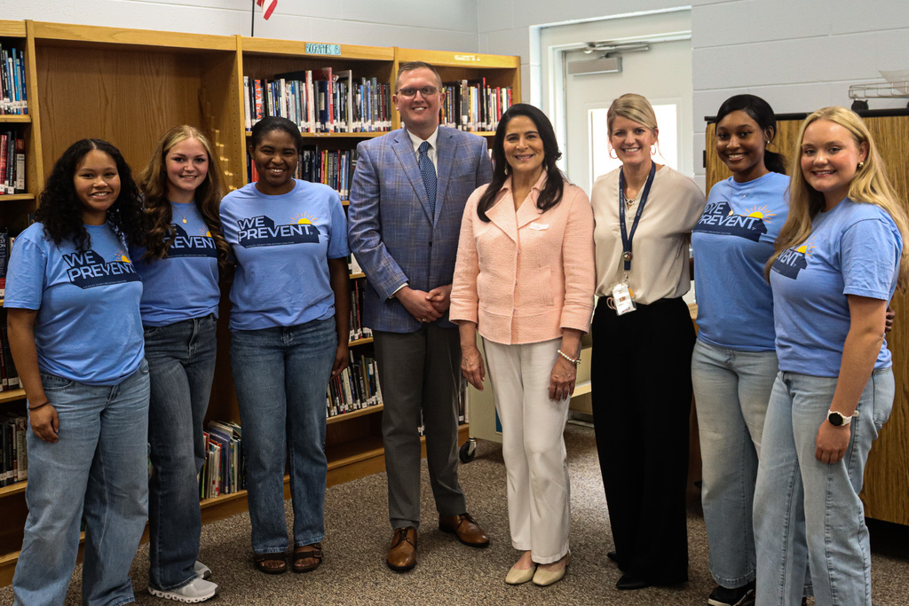 Nine people stand in a room, smiling for a photo. Two men in suits and ties, six women in jeans.