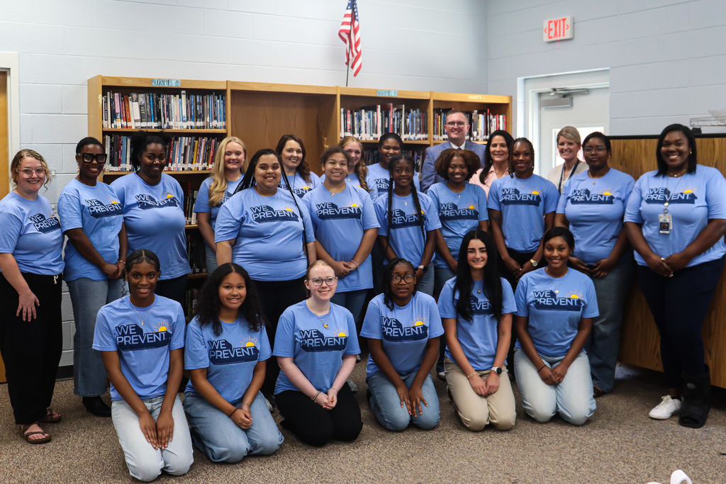 Group in blue shirts posing for photo in room with bookshelves and flag, some seated on floor.