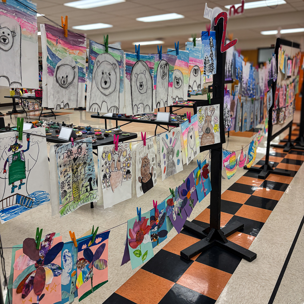 A room with artwork displayed on a clothesline, tables, and floor, illuminated by fluorescent lights.