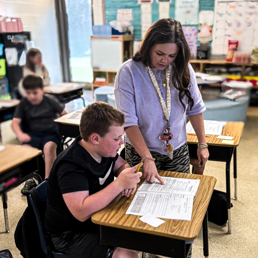 A teacher assists a student at a desk, with other students in the background in a classroom.