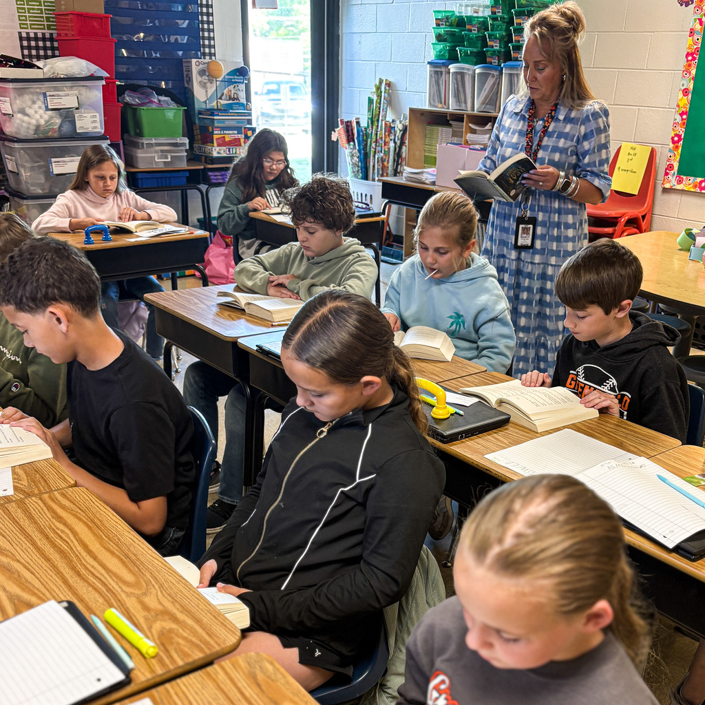 A classroom with students seated at desks, reading books. A teacher stands behind them, reading from a book.