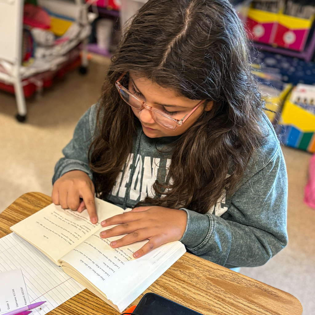 A girl with glasses and a gray sweatshirt reads a book in a classroom setting.