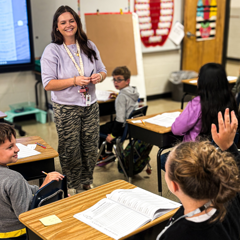 A teacher stands in front of a classroom. Students sit at desks, one raising a hand. A board and trash can are in the background.