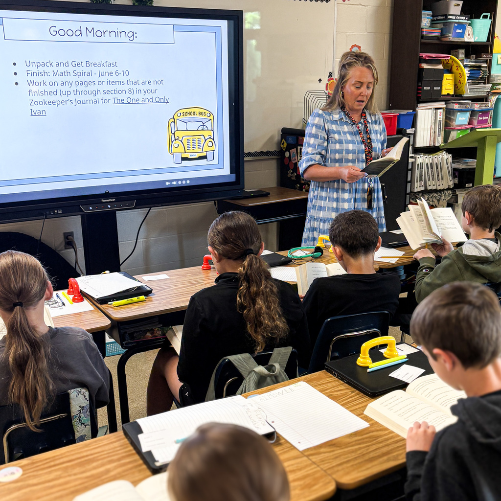 A classroom with students seated at desks, a teacher reading from a book at the front.
