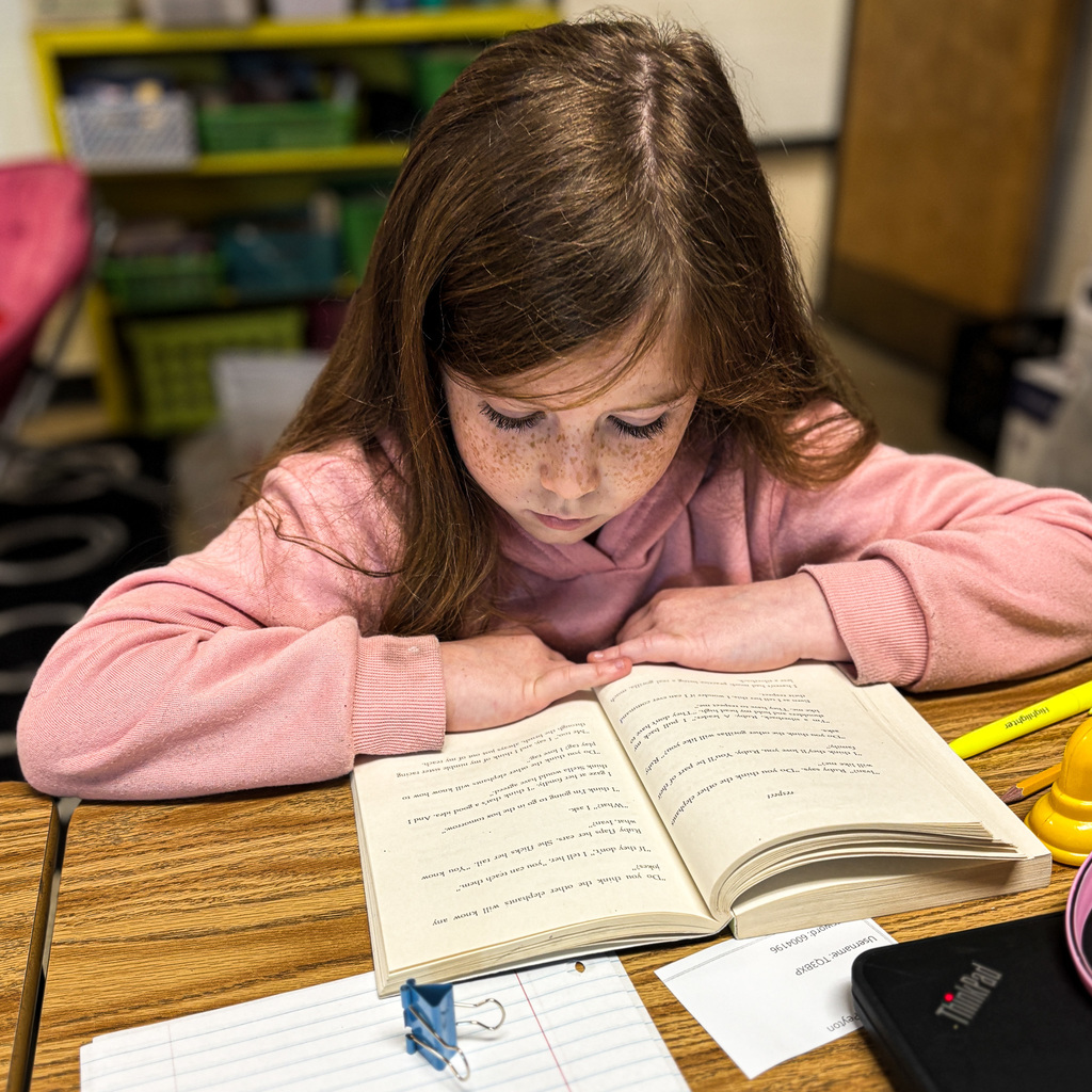 A girl with brown hair reading a book at a table, surrounded by papers and a yellow pencil.
