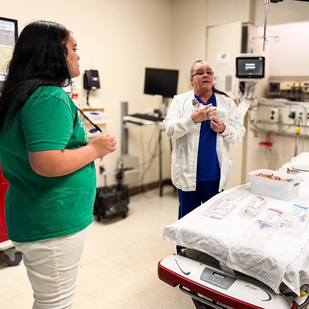 Two women stand in a hospital room. One wears green; the other, white. A table with medical items is nearby.