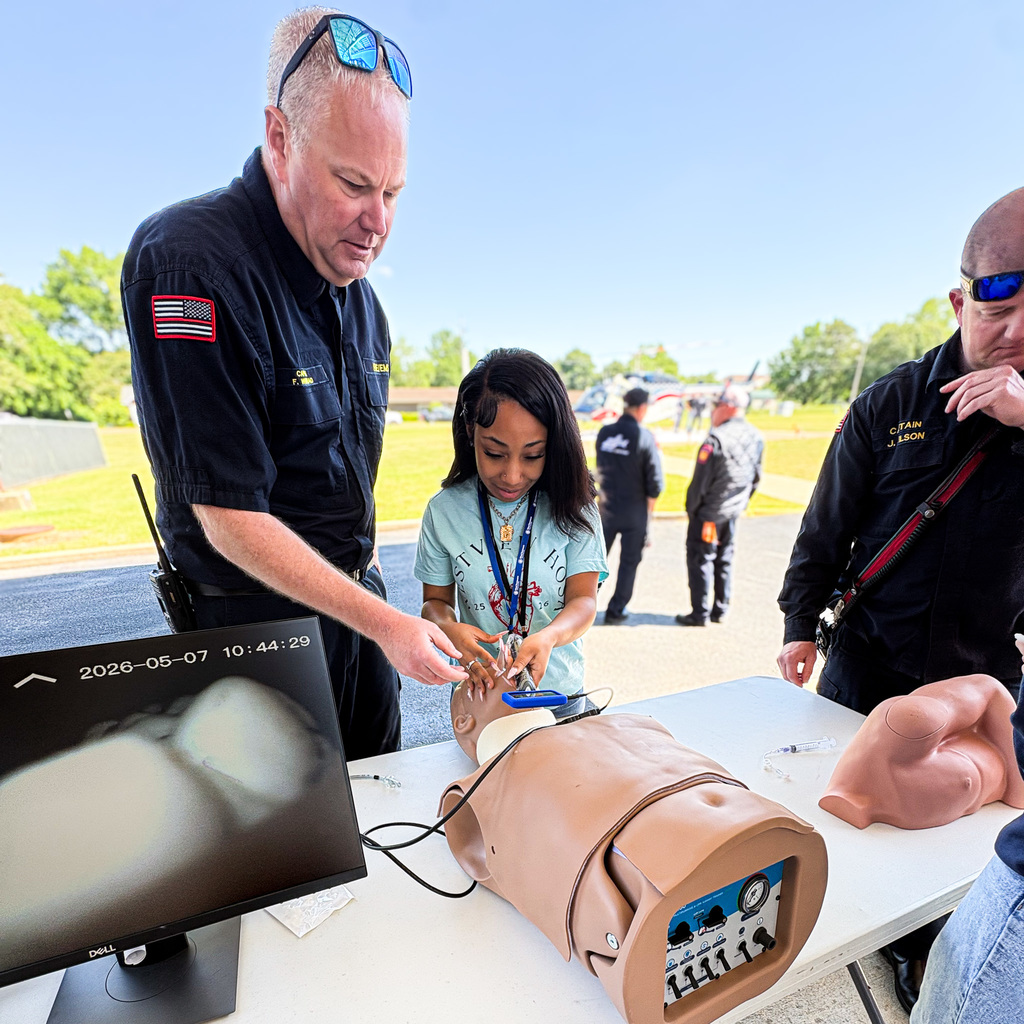 A person adjusts a monitor while another person operates a dummy on a table. Behind them, others observe.