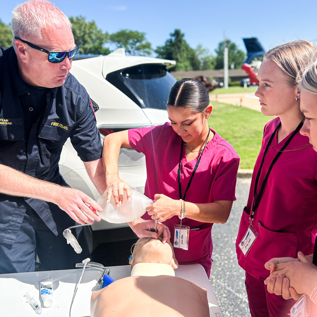 Three women and a man practice first aid on a dummy, outdoors with a white car in the background.
