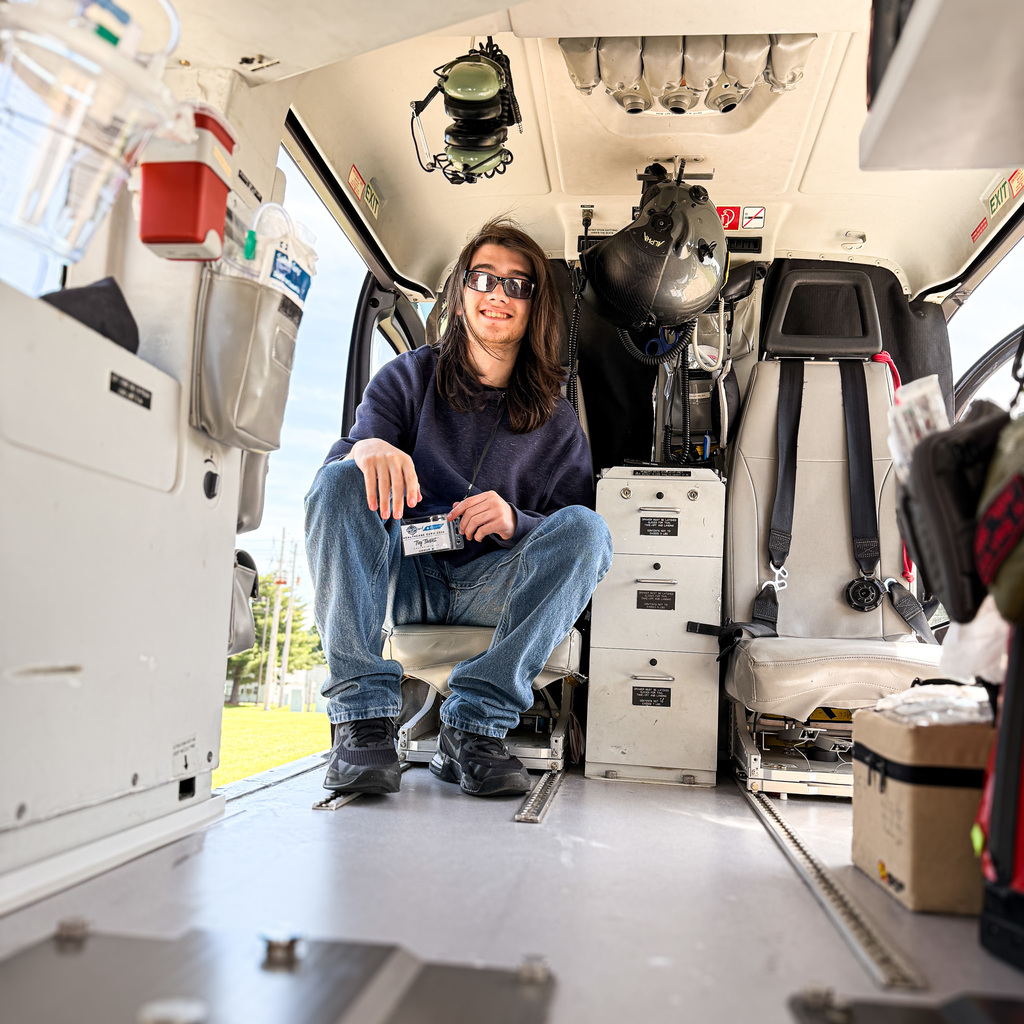 Person sitting on chair inside helicopter, wearing sunglasses, holding a badge, and looking at camera.