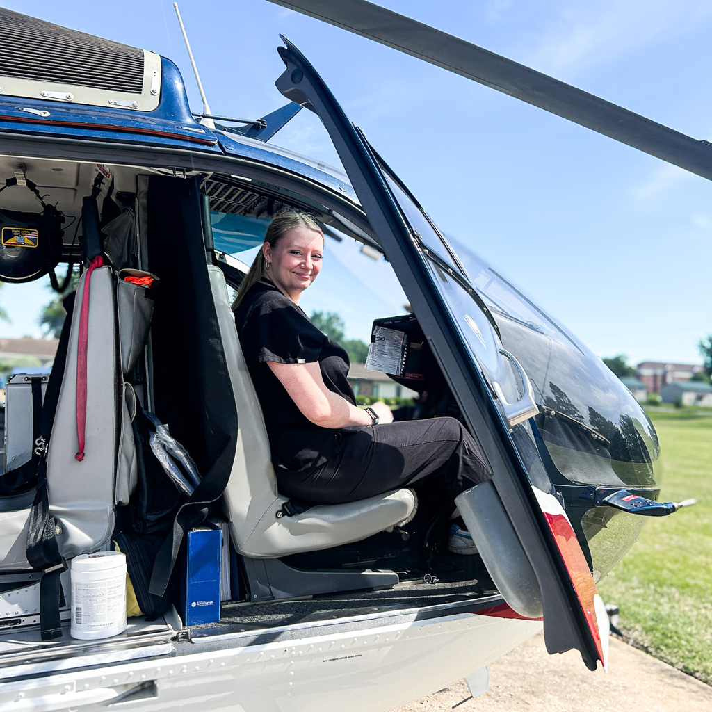 A person sits in the cockpit of a helicopter, smiling. The helicopter door is open, revealing the cockpit's interior.