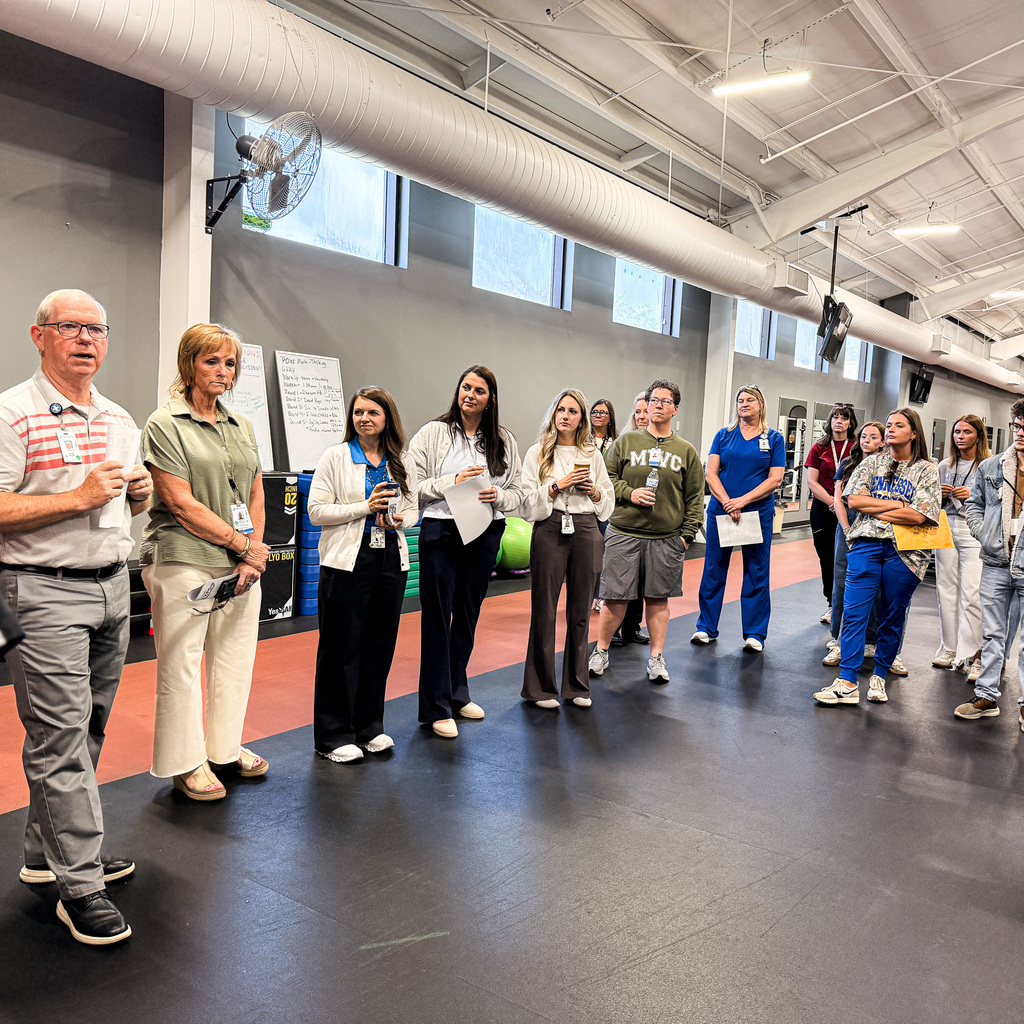 A group of people in matching uniforms stand in a line in a gym with white pipes.