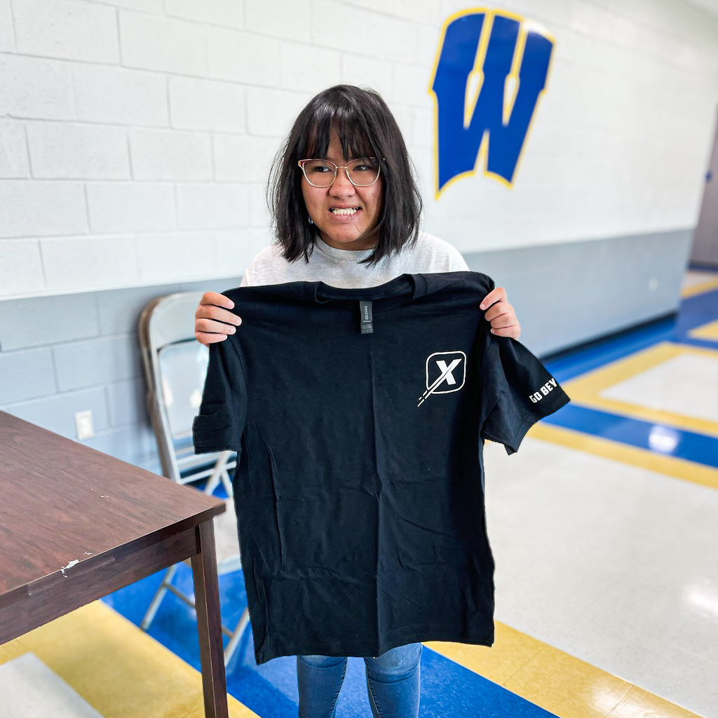 A person holds a black t-shirt with a white logo in a room with white walls and a blue floor.