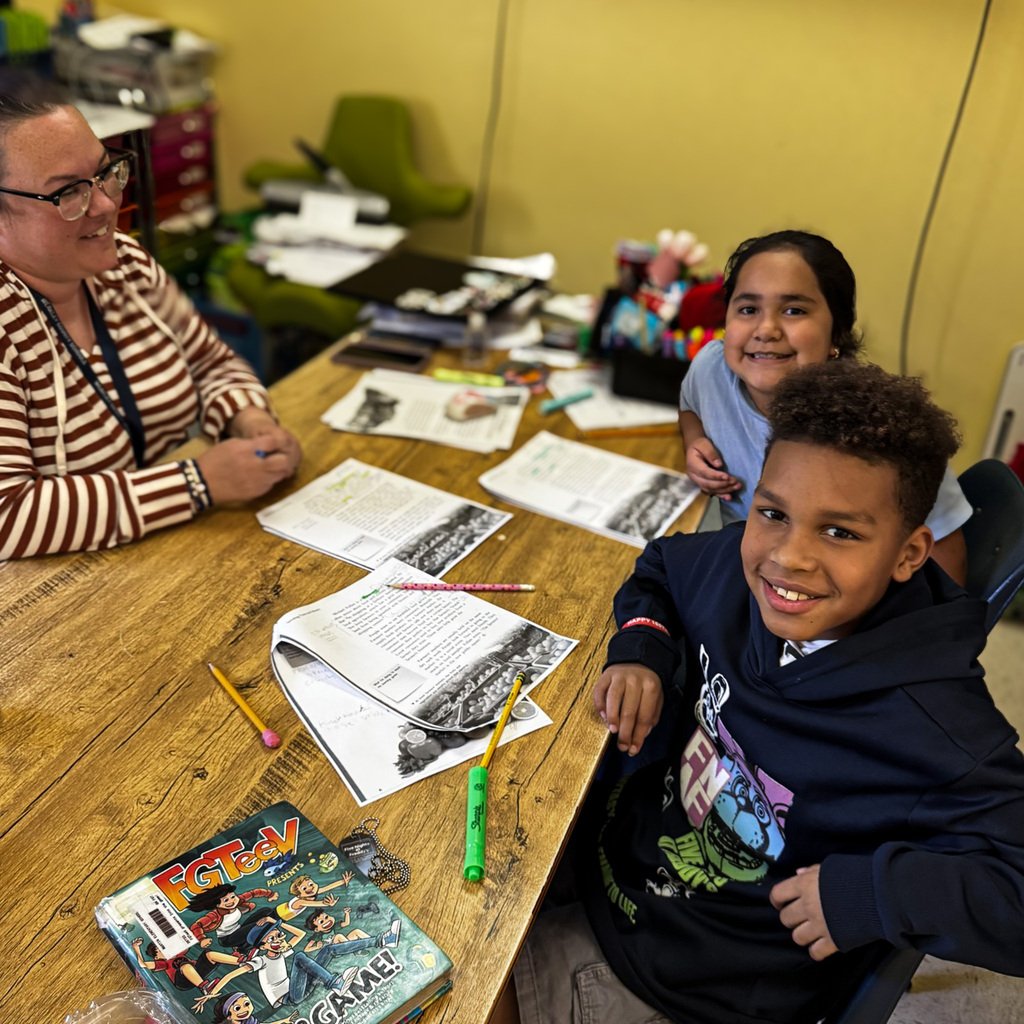 A woman and two children sit at a desk with papers, pencils, and a book.