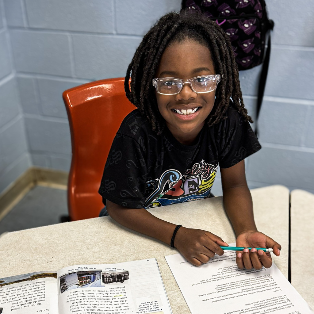 A child with glasses sits at a desk, smiling, holding a pencil, and writing on a paper.