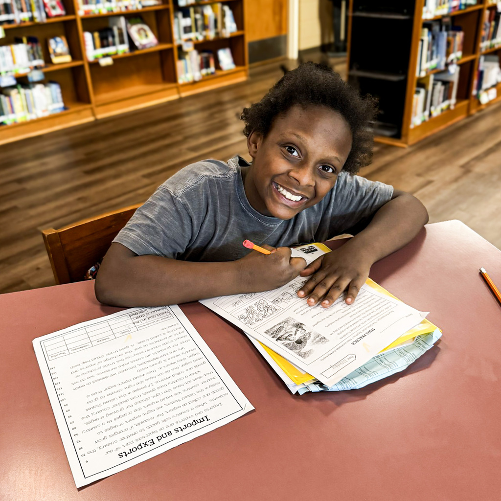 A child seated at a table, smiling while holding a pencil and working on a worksheet.